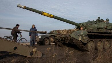 Ukrainian serviceman looks on and a local resident rides a bicycle while a broken tank is pulled to a truck near the frontline town of Bakhmut