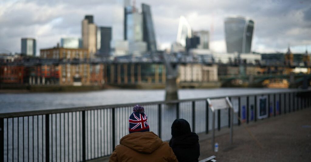 People sit alongside the bank of the River Thames with the City of London financial district in the background, in London